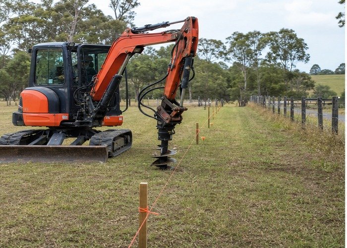 Mini excavator with auger aligned beside a marked fence line