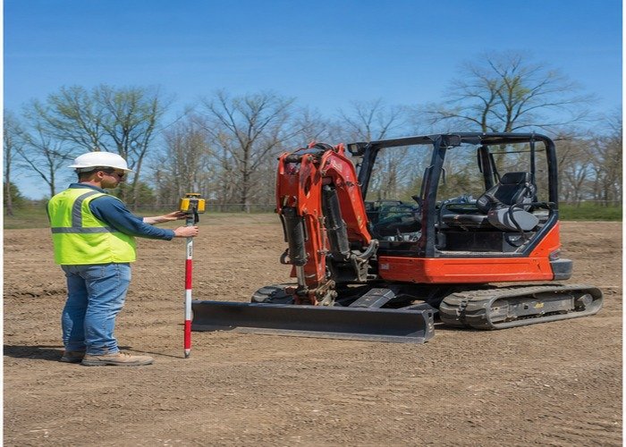 Operator checking a grade rod beside a stopped mini excavator