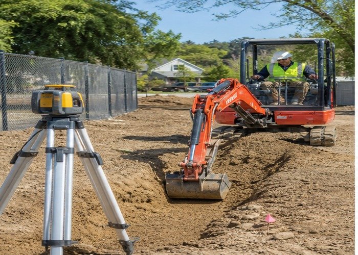 Mini excavator cutting a shallow trench beside a safely positioned rotating laser
