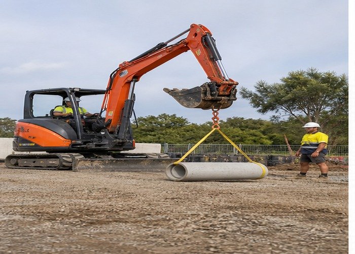 Mini excavator lifting a short pipe section close to the ground with rated rigging