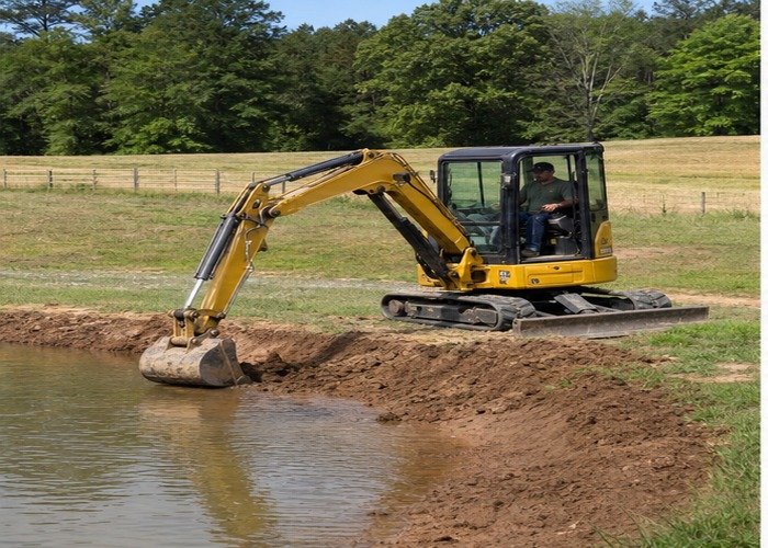 Mini excavator shaping a small farm pond edge from stable ground