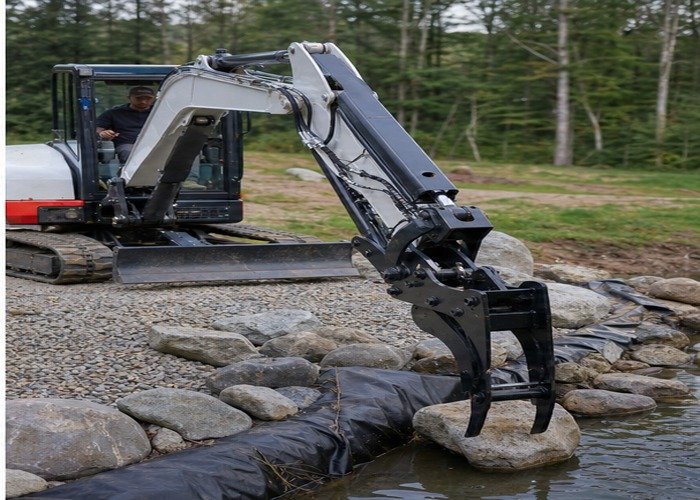 Mini excavator hydraulic thumb placing rocks near drainage fabric with a clear work zone