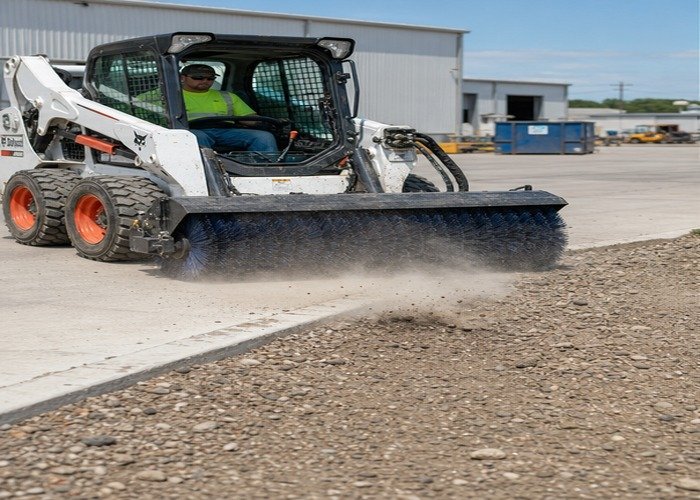Skid steer rotary broom sweeping gravel away from a concrete apron