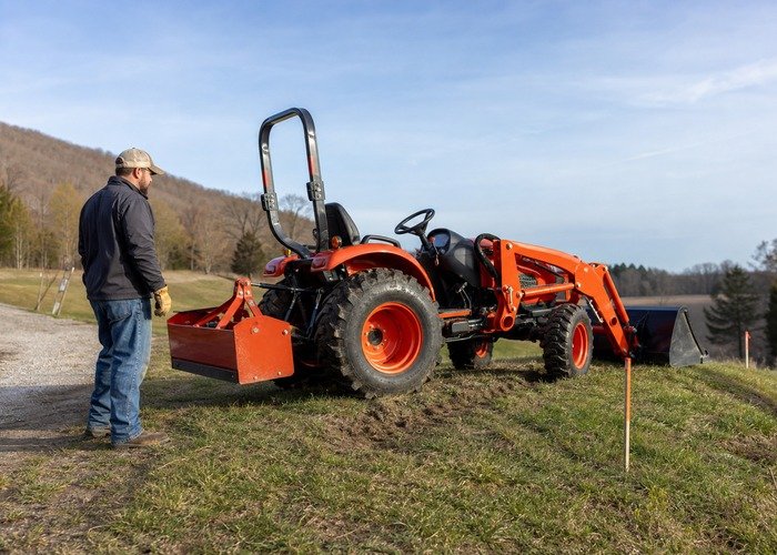 Tractor ballast and slope route check Compact tractor on gentle rural ground with low loader bucket and rear ballast while an operator checks the route