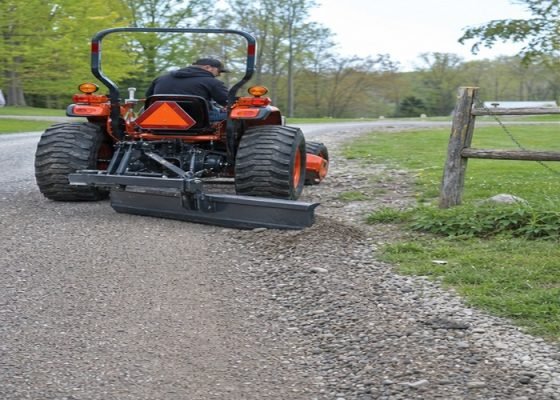 Compact tractor rear blade pulling loose gravel from driveway shoulder back toward the lane
